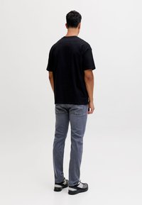 Mand iført sort T-shirt med korte ærmer, grå jeans og sort-hvide sneakers, stående med ryggen modskærmen mod en ensartet lys baggrund.