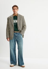 Blazer gris de cuadros sobre un sudadera verde, jeans de pierna ancha azul claro y zapatos beige. Corte recto con detalles de costura visibles.