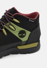 Scarpa da trekking nera e verde con tomaia in rete, suola in gomma e dettagli testurizzati. Presenta il logo e una fodera interna rossa a contrasto.