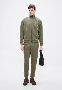 Σετ φόρμας σε ελιά πράσινο με τζάκετ που κλείνει με φερμουάρ και παντελόνι ταιριαστό, με αντίθετες ρίγες, ελαστικές μανσέτες και μικρό λογότυπο. Μαύρη τσάντα στο χέρι.