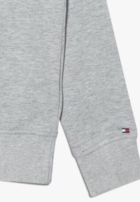 Felpa grigia con polsini e orlo a coste. Presenta una piccola patch del logo in rosso, bianco e blu. Tessuto morbido, design casual.