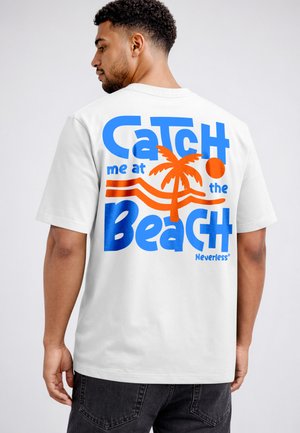 Mann trägt weißes T-Shirt mit blauem und orangem Text und Grafiken mit der Aufschrift „Catch me at the beach“ und einem Palmen- und Sonnendesign.