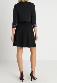 Černé šaty s přiléhavým živůtkem a rozšířenou sukní. Tříčtvrteční rukávy zdobí květinové flitrové detaily na manžetách. Nosit s černými vysokými podpatky.