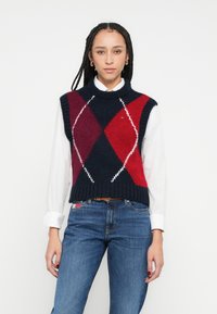 Pletená bezrukávová vesta s námořnicky modrým, červeným a bielym vzorom argyle; rebrovaný golier a lem; nosená cez bielu košeľu s dlhým rukávom.