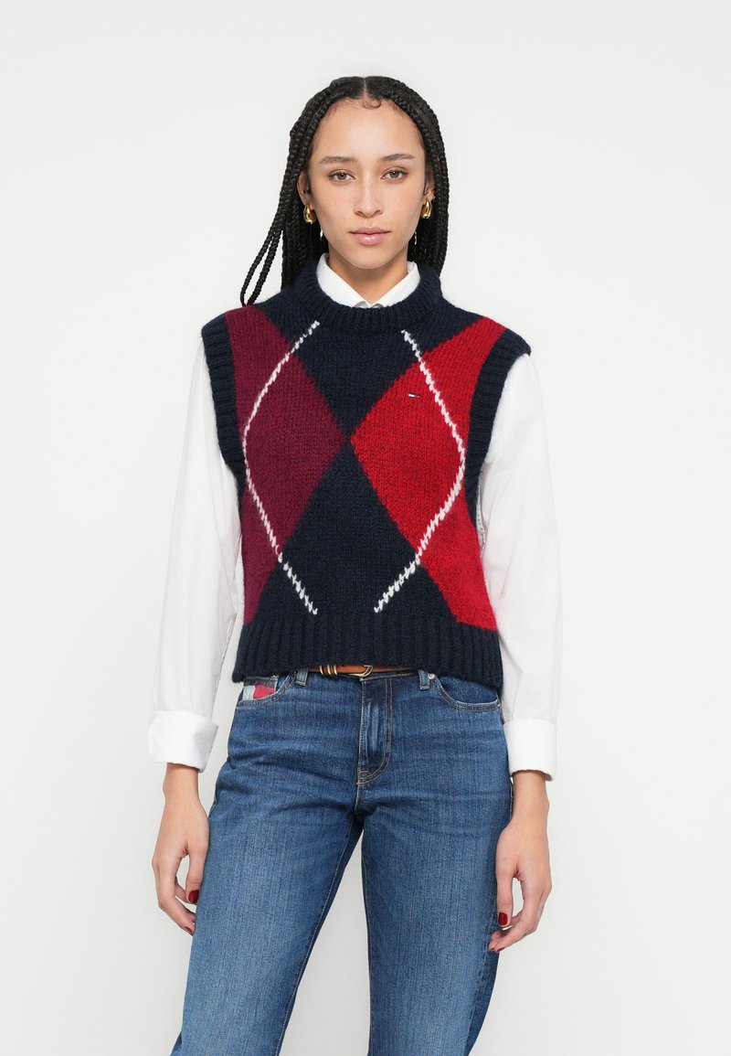 Pletená bezrukávová vesta s námořnicky modrým, červeným a bielym vzorom argyle; rebrovaný golier a lem; nosená cez bielu košeľu s dlhým rukávom.
