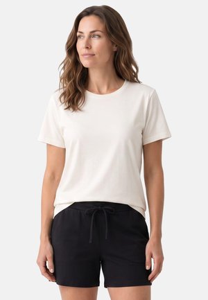 Femme aux longs cheveux bruns portant un t-shirt blanc uni à manches courtes et un short noir à cordon, regardant vers la gauche sur un fond neutre.