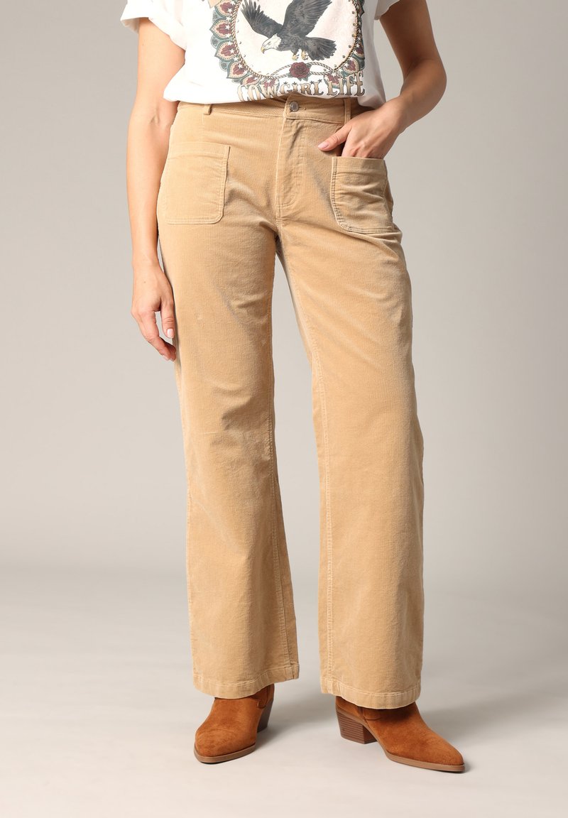Pantalons en velours côtelé beige à jambes larges, avec deux poches avant. Associés à des bottes à talons en daim marron et un t-shirt graphique.
