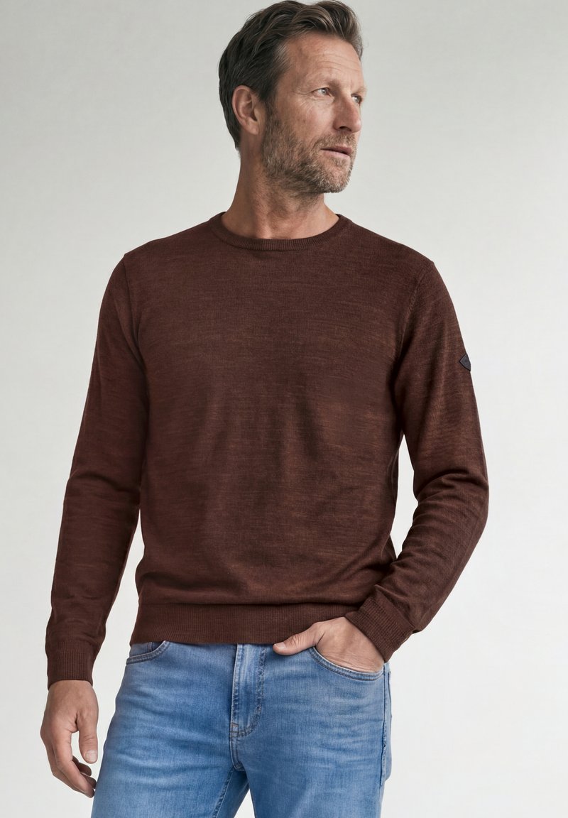 Homme d'âge moyen aux cheveux courts et à la barbe, portant un pull marron et un jean bleu, regardant vers la droite, avec une main dans la poche.