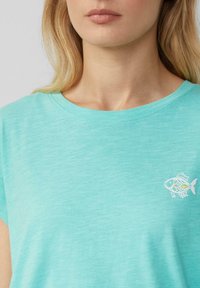 Vrouw in een lichtblauwe shirt met ronde hals, met een klein wit en geel visje op de linkerkant van de borst.