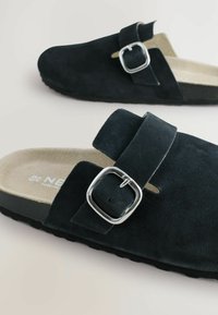 Crne nubuk sandale na vezivanje bez kopčanja s širokim remenom, detaljem srebrne kopče i lagano podstavljenim bež uloškom, koje se odmaraju na teksturiranoj crnoj potplati.