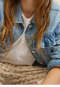 Džínová bunda se žabkovaným límcem, světle modrá barva, nošená přes bílou košili. Vzorovaná květinová látka vykukuje z béžových kalhot.