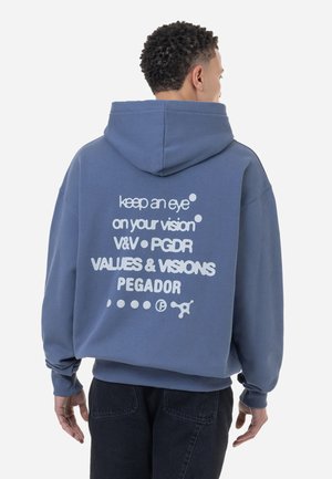 Oversized blue hoodie with a soft texture. White text on the back includes various phrases and brand names. Features a spacious hood.