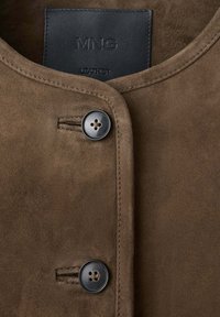 Veste en daim marron avec un col rond, deux boutons noirs et des coutures visibles autour du col et des boutonnières.