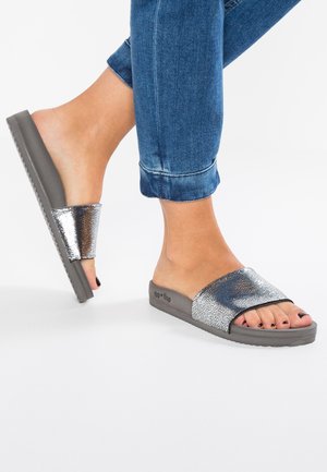 Silver textured slide sandals with a wide strap, gray sole, open toe, and a flat design, paired with rolled-up denim jeans.