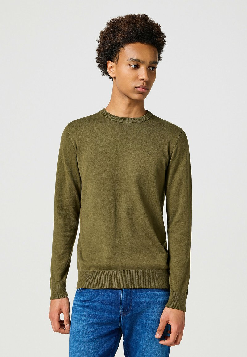 Maglione a maglia verde oliva con scollatura rotonda, maniche lunghe e polsini a coste. Indossato su jeans blu, presenta un design semplice e strutturato.