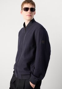 Marineblauwe fleece bomberjack met een voorrits, geribde manchetten en een logo-opdruk op de mouw. Relaxte pasvorm met een zachte textuur.