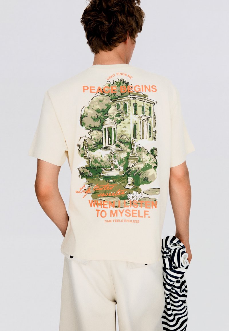 Homme portant un t-shirt crème avec une illustration de jardin vert et statues classiques et texte orange au dos. Homme tient un vêtement rayé noir et blanc.