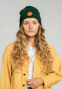 Zielona dziana czapka z okrągłą skórzaną łatą w kolorze brązowym. Nosi ją z żółtą kurtką dżinsową na białej koszuli, prezentując swobodny, warstwowy wygląd.