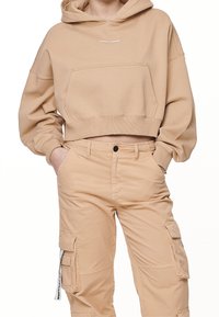 Hoodie recortado beige con un bolsillo frontal y capucha, que presenta un logo blanco. Combinado con pantalones cargo de color beige claro con bolsillos laterales.