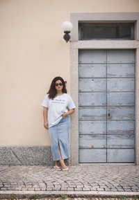 T-shirt graphique bleu clair avec texte, associé à une longue jupe en denim avec une fente à l'avant. Sandales blanches, se tenant devant un mur texturé.