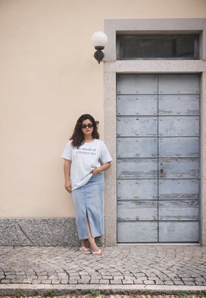 Camiseta gráfica azul claro con texto, combinada con una falda larga de mezclilla con abertura frontal. Sandalias blancas, de pie contra una pared texturizada.