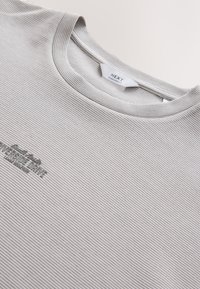 Lys gråstribet t-shirt i oversized pasform med rund halsudskæring og diskret broderet logo på brystet. Blød tekstur og afslappet design.