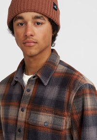 Flonel skjorte i brune, grå og orange tern med trykknapper og en brystlomme. Modellen bærer en brun strikket beanie og en halskæde.