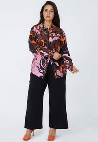Woman adjusting cuff of colorful patterned long-sleeve shirt, wearing black wide-leg pants and orange high-heeled shoes, standing on white background.