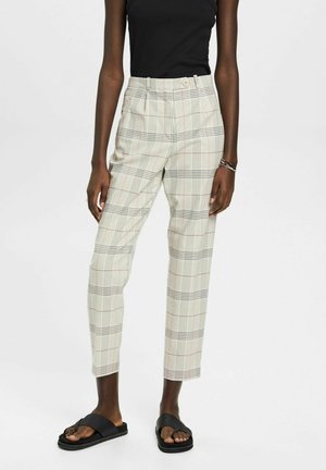 Osoba nosząca beżowe, kraciaste spodnie o krótkiej długości z wysokim stanem i czarne klapki, w połączeniu z czarnym, bezrękawnym topem.