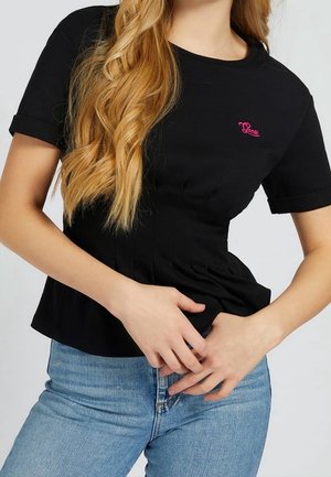 Kvinde iført sort kortærmet skjorte med pink Guess-logo og blå jeans, med langt bølget blondt hår over skulderen.