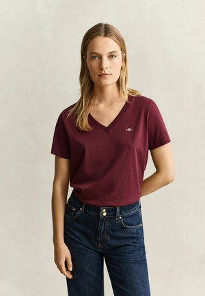 Frau mit schulterlangem blondem Haar, die ein bordeauxrotes T-Shirt mit V-Ausschnitt und dunkelblaue Jeans trägt, vor einem schlichten hellen Hintergrund stehend.