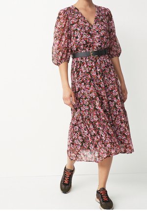 Femme portant une robe midi fleurie rose et rouge avec des manches bouffantes, une ceinture noire et des baskets vert foncé avec des lacets orange, marchant vers l'avant.