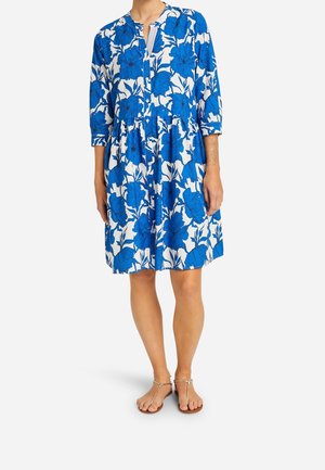 Blau-weißes Blumenmuster-Kleid mit 3/4-Ärmeln, geraffter Taille und Schlitzdetail am Ausschnitt. Kombiniert mit flachen Riemchensandalen.