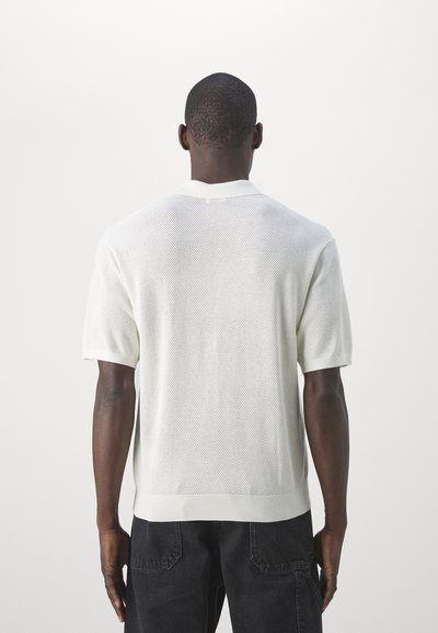 Polo shirt tricot à manches courtes blanches avec un design en maille, un col côtelé et un ourlet. Porté à l'envers, sur un fond neutre.
