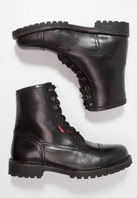 Bottes basses en cuir noir avec bout rond, semelle plate et laçage à l'avant. Elles présentent une étiquette logo rouge et une semelle extérieure texturée et robuste.