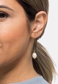 Gullkjedeøredobber med en hengende, oval hvit perle. Designet inkluderer små gulldetaljer som gir et elegant, minimalistisk utseende.