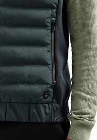 Gevoerde vest in donkergroen, met een ritssluiting aan de zijkant, geribbelde zoom en een textuur groene mouw. Het materiaal is glad met een contrasterende brei.