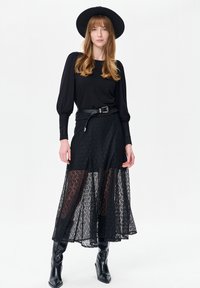 Zwarte blouse met opgeblazen mouwen, gecombineerd met een doorzichtige zwarte kanten rok met bloemmotieven, en zwarte kniehoge laarzen. Zwarte hoed met brede rand.