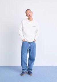 Fehér, hosszú ujjú ing, apró mellnyomattal, laza szabású világoskék farmernadrággal és sárga talpú szürke cipővel párosítva.