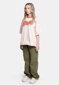 Beige oversized t-skjorte med rød krage og oransje droppende grafikk; kombinert med olivengrønne baggy bukser og kremfargede sko.