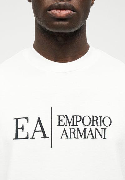 Κοντινό πλάνο ενός ατόμου που φοράει λευκό πουκάμισο με κεντημένο μαύρο λογότυπο "EA | Emporio Armani" στο στήθος.
