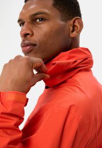 Rode waterdichte jas met hoge kraag en verstelbare manchetten. Gestructureerde stof en zwarte hardware details voor een veilige sluiting.