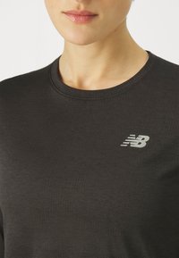 Musta urheilullinen pitkähihainen paita, jossa on uritettu pinta, ja vasemmassa yläosassa hopea New Balance -logo. Pyöreä kaula-aukko.
