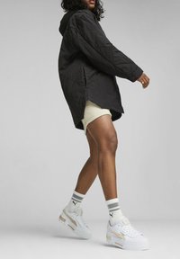 Chaqueta larga acolchada negra con capucha, con aberturas laterales, llevada sobre una falda de canalé crema y deportivas blancas gruesas con acentos dorados.