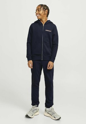 Felpa con zip di colore navy, dotata di tasche frontali, abbinata a pantaloni coordinati. Presenta un logo sul petto e polsini a costine. Indossata con sneakers nei toni neutri.