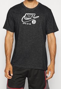 Hombre vistiendo una camiseta de Nike color gris oscuro con logo de baloncesto de los Brooklyn Nets y pantalones cortos de malla negros con ribete rojo, torso visible.