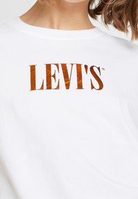 Valkoinen puuvillateeppari, jossa on brändin nimi "LEVI'S" suurilla, metallinhohtoisilla kuparinvärisillä kirjaimilla rinnan kohdalla. Sujuva rakenne.