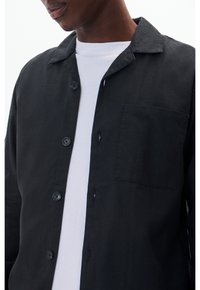 Czarna koszula zapinana na guziki z kołnierzykiem, z jedną naszywaną kieszonką oraz widoczną fakturą materiału. Nałożona na białą koszulkę.