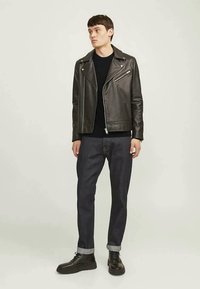 Homem vestindo uma jaqueta de motociclista de couro preta, um suéter preto, jeans escuros com as bainhas enroladas e botas de couro pretas, encostado a um fundo claro e simples.