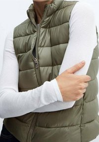 Zielono-oliwkowa puchowa kamizelka z wysokim kołnierzem, zapinana na zamek, o pikowanej strukturze, noszona na białej bluzce z długim rękawem.